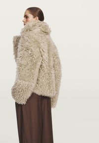 Faux-Fur-Jacke in hellem Beige, mit fluffiger Textur, weiten Ärmeln und hohem Kragen, kombiniert mit dunkelbrownen weiten Hosen.