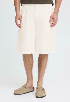 Pantalones cortos hasta la rodilla de color crema, hechos de algodón, con un pliegue frontal y bolsillos laterales. Combinados con zuecos de ante color canela.