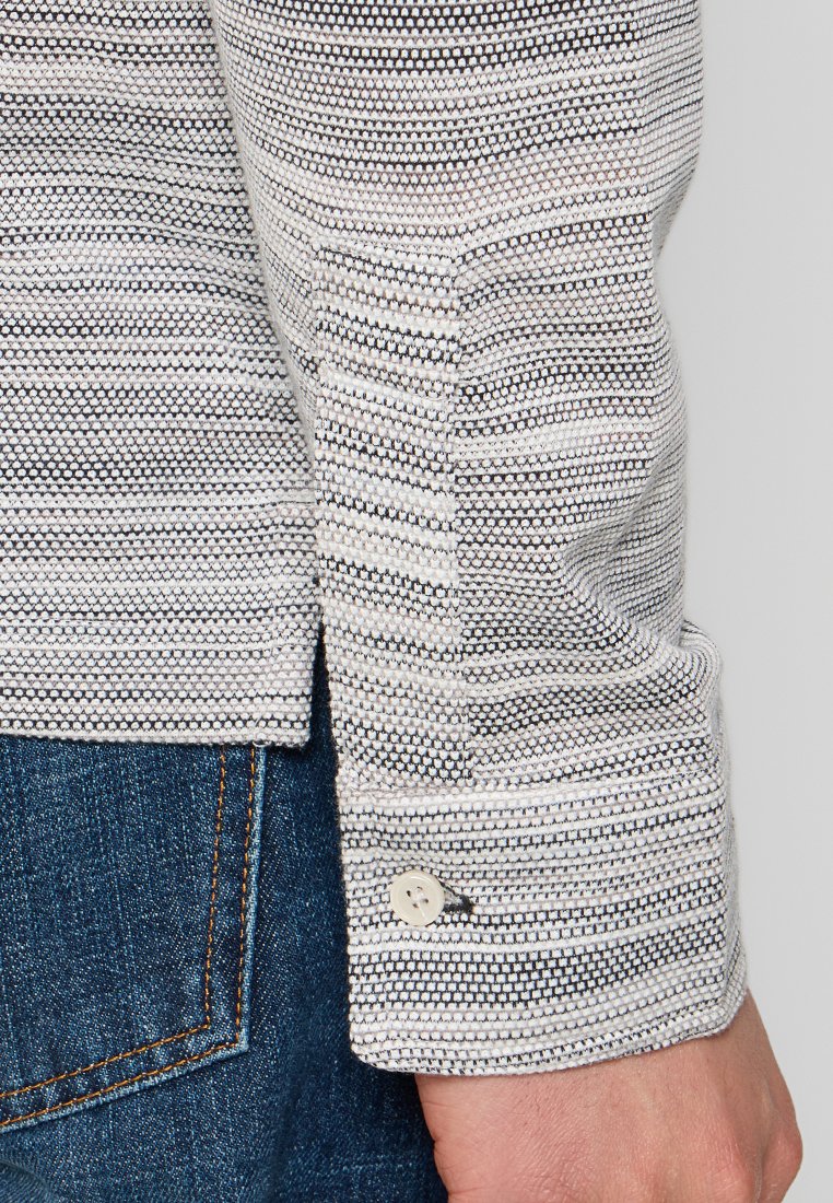 Gestreiftes Baumwollhemd mit Mustern in Schwarz, Weiß und Grau. Mit einem Knopf versehene Manschetten und ein gerader Saum. Hintergrund aus blauer Denim.