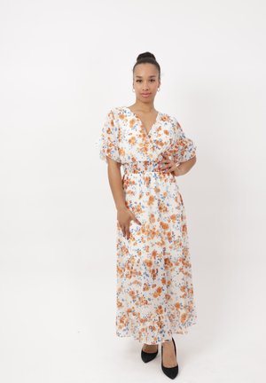 Kvinde med håret i en knold iført en hvid blomster maxi-kjole med orange og blå blomster, poserer med den ene hånd på hoften og sorte hæle.
