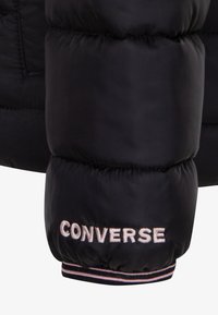 Veste matelassée noire avec un design matelassé, des poignets élastiques et "CONVERSE" brodé en fil clair sur la manche.