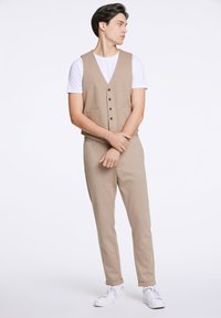Beige vest met knopen en voorzakken, gecombineerd met bijpassende pantalon. Gedragen over een wit t-shirt, afgewerkt met witte sneakers.
