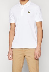 Polo blanc en coton, avec un col pointu et trois boutons. Un petit logo jaune est brodé sur le côté gauche de la poitrine.