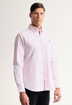 Camisa de botones a rayas en rosa claro con mangas largas, cuello y un pequeño logotipo bordado en el pecho. Usada con pantalones negros.