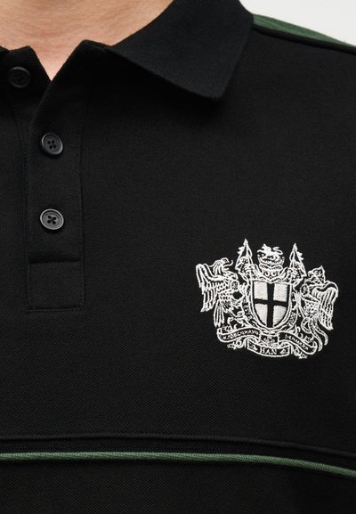 Μαύρο πόλο πουκάμισο με τρία μαύρα κουμπιά, μοντέρνο λευκό κεντητό έμβλημα που απεικονίζει ασπίδα και δύο ερμηνευτικά ζώα, και μία λεπτή πράσινη ρίγα.