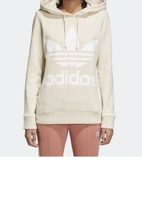 Béžová mikina s velkým bílým logem Adidas trefoil, kapucí na šňůrku a žebrovanými manžetami. V kombinaci se světle růžovými tepláky se bílými pruhy.