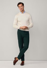Ein hellcremer Pullover über einem hellblauen Hemd, kombiniert mit dunkelgrünen Hosen und braunen Lederschuhen. Das Outfit hat ein lässiges, eng geschnittenes Design.
