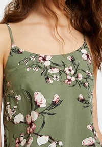 Bezrękawnik w oliwkowym kolorze, w kwiatowy wzór, z różowymi i białymi kwiatami oraz czarnymi akcentami, charakteryzujący się prostym, luźnym krojem.