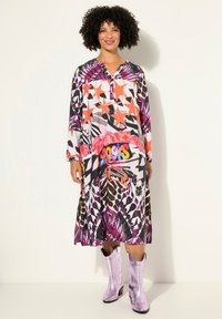 Langes, farbenfrohes Kleid mit auffälligen Mustern in Lila, Pink und Orange. Es hat einen Henley-Ausschnitt und wird mit glänzenden, helllila Stiefeln kombiniert.