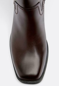 Tmavohnedá kožená čižma so štvorcovou špičkou a čiernou podrážkou, ktorá ukazuje hladkú textúru a vrchné prešívanie blízko členka.