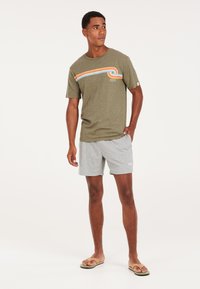 Camiseta verde olivo con rayas horizontales multicolor, combinada con pantalones cortos grises. Modelo usando sandalias, de pie sobre un fondo claro.