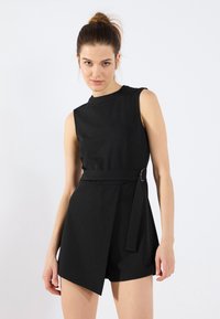 Combinaison sans manches noire en tissu lisse, avec un décolleté arrondi, une taille ceinturée dotée d'une boucle circulaire et des shorts asymétriques.