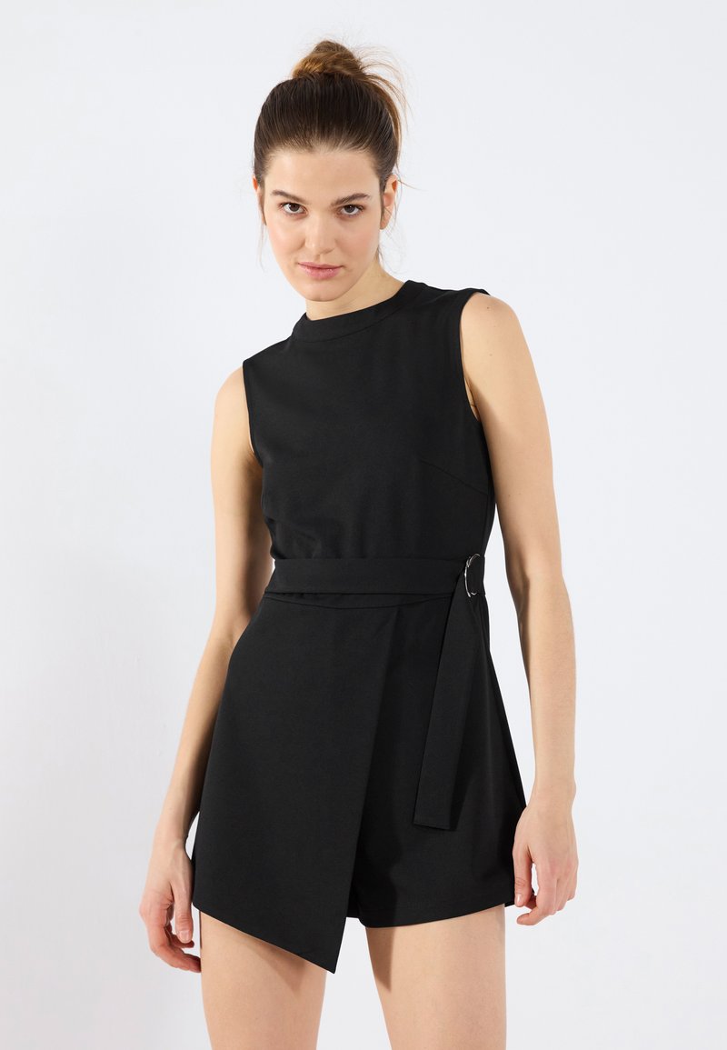 Combinaison sans manches noire en tissu lisse, avec un décolleté arrondi, une taille ceinturée dotée d'une boucle circulaire et des shorts asymétriques.