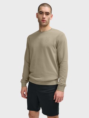 Man met kort haar, gekleed in een beige sweatshirt met lange mouwen en zwarte korte broek, staand tegen een effen lichtgrijze achtergrond.