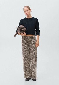 Črna pletena kratka jopica z kabelskim vzorcem, kombinirana s širokimi tan hlačami z leopardjim potiskom. Model drži rjav nahrbtnik.