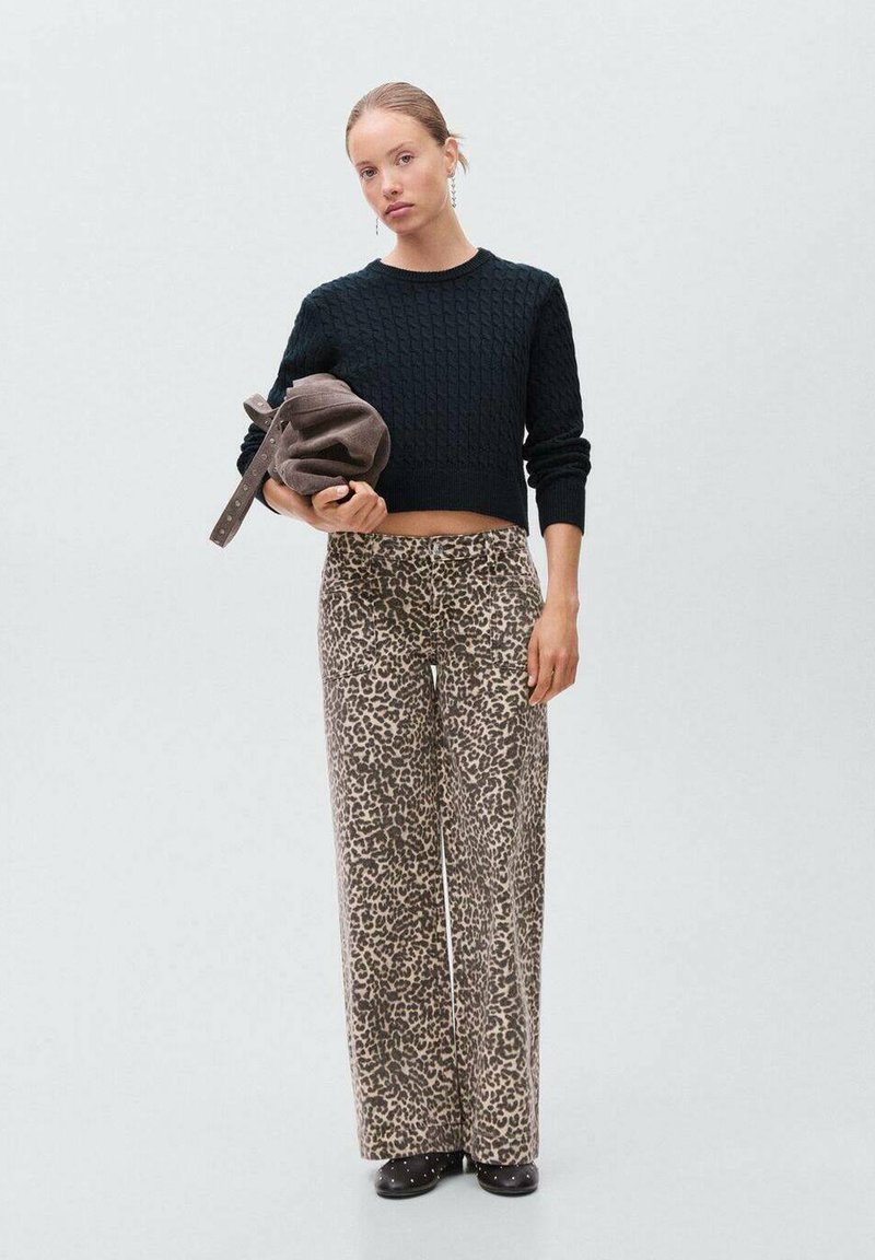 Črna pletena kratka jopica z kabelskim vzorcem, kombinirana s širokimi tan hlačami z leopardjim potiskom. Model drži rjav nahrbtnik.