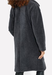 Donna che indossa un lungo cappotto in pile sherpa grigio scuro con maniche lunghe, in piedi con la schiena rivolta verso la macchina fotografica.