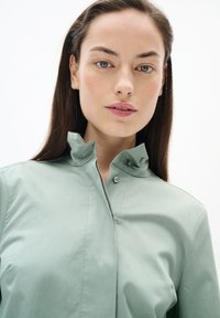 Hellgrünes Hemd mit durchgehendem Knopfverschluss, hochgeschlossenem Kragen, glattem Textil und dezenten Nahtdetails. Der Stoff wirkt leicht und atmungsaktiv.