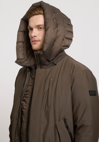 Veste matelassée brune avec une grande capuche repliée, fermeture éclair frontale et poches latérales. Fabriquée en tissu léger et lisse avec des détails texturés.