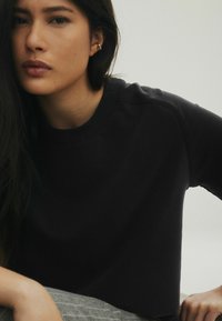 Schwarzes Baumwoll-Sweatshirt mit rundem Ausschnitt und lockerer Passform, ausgestattet mit gerippten Bündchen und einer glatten Textur. Der Hintergrund ist hellgrau.