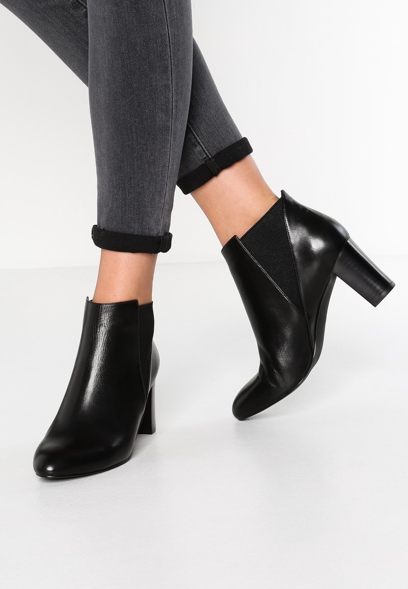 Bottes chevilles en cuir noir avec talons carrés portées avec un jean skinny gris foncé retroussé sur des jambes croisées.