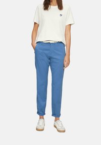Jasno beżowa koszulka z wzorem serca, zestawiona z niebieskimi chinosami i białymi trampkami na gumowej podeszwie. Chinosy mają podwinięte nogawki i kieszenie.