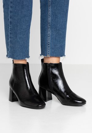 Bottines en cuir verni noir brillant à talons blocs portées avec un jean bleu court à ourlet effiloché sur un sol clair.