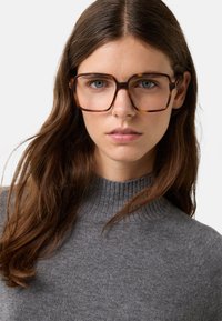 Ung kvinde med langt brunt hår iført store brune skildpaddeskalsbriller og en grå ribbet turtleneck sweater, neutral baggrund.