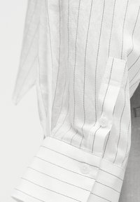 Hvit langermet skjorte med mansjett og erme, tynne vertikale svarte nålestriper og hvite knapper på mansjett og ermeliste.