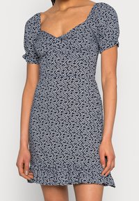 Marinblå klänning med vitt blommönster, korta puffärmar och fyrkantig halsringning. Har en volang i nederkant och en figurnära design.