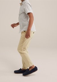 Camicia bianca a maniche corte con macchie scure, pantaloni beige e mocassini blu navy. Dettaglio del papillon visibile sul colletto della camicia.