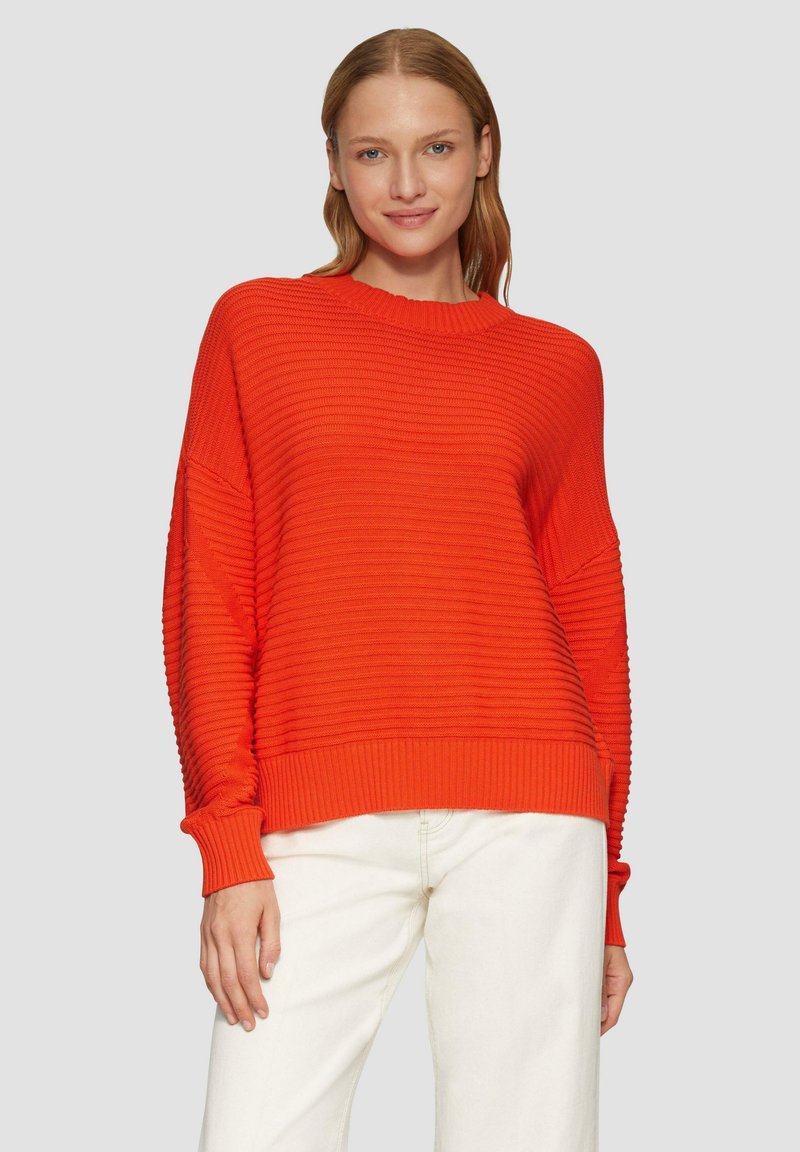 Roter, gerippter Strickpullover mit Rundhalsausschnitt und lockerer Passform. Lange Ärmel mit gerippten Bündchen, kombiniert mit hellen Hosen.