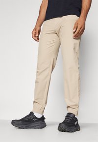 Beige lehké kalhoty se slim fit a elastickými manžetami, spárované s černými sportovními botami s texturovaným designem a viditelným logem.