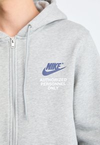 Siva zip-up kapuca iz mehkega materiala, ki ima modri Nike logotip in belo besedilo, ki pravi "DOVOLJENI SAMO OBLASTI".