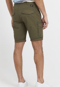 Shorts cargo vert olive avec ourlets roulés, texture subtile, plusieurs poches et une étiquette à la taille. Portés avec des baskets blanches.