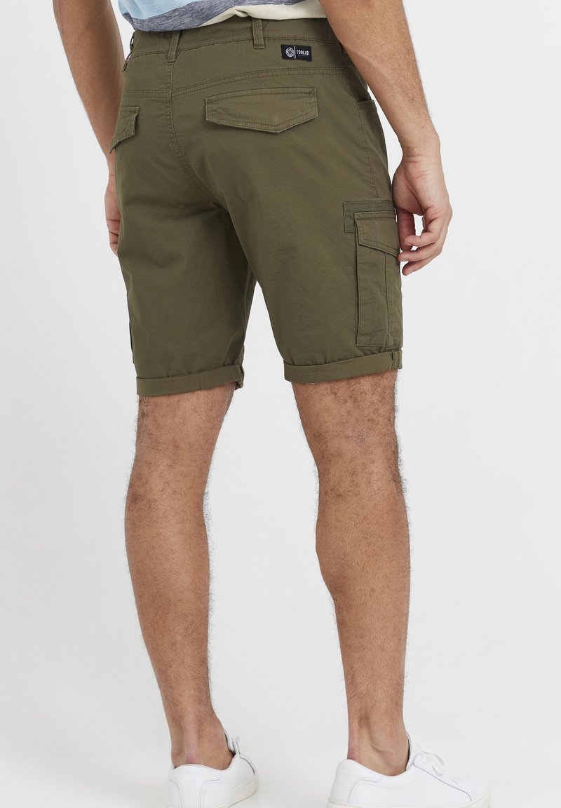 Shorts cargo vert olive avec ourlets roulés, texture subtile, plusieurs poches et une étiquette à la taille. Portés avec des baskets blanches.