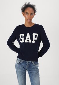Marineblauer Strickpullover mit großen, cremefarbenen "GAP"-Buchstaben auf der Vorderseite. Kombiniert mit hellblauen Jeans. einfacher runder Halsausschnitt und langen Ärmeln.