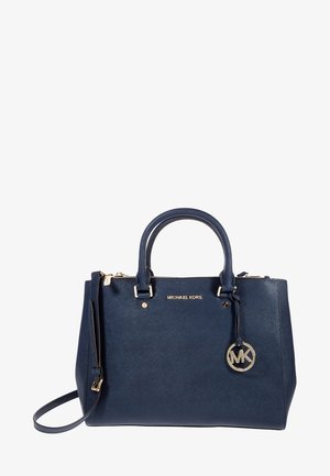 Sac à main Michael Kors bleu foncé avec deux anses, une bandoulière, une plaque logo dorée et un pendentif rond MK suspendu sur le côté.