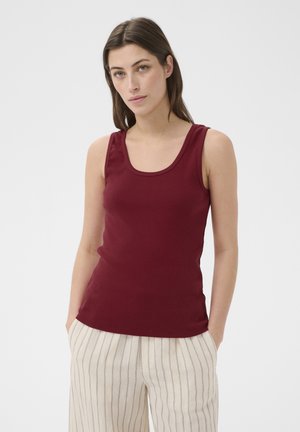 Femme aux cheveux bruns lisses portant un haut bordeaux sans manches et un pantalon blanc à rayures verticales, les mains dans les poches, sur un fond uni.