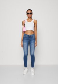 Žena s čiernymi slnečnými okuliarmi, bielym krátkym tielkom, modrými úzkymi džínami, bielymi teniskami, náušnicami v tvare kruhu a malou ružovou kabelkou cez plece.