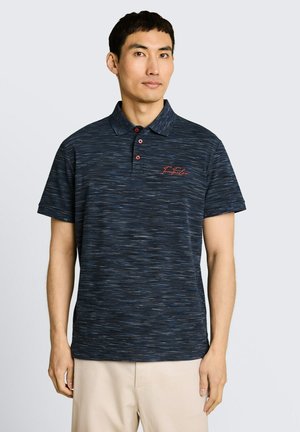 Homme portant un polo bleu foncé à motif avec des boutons rouges et un logo, associé à un pantalon beige clair, se tenant devant un fond uni.