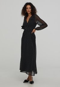 Czarna sukienka maxi w białe kropki z długimi prześwitującymi rękawami, w stylu kopertowym, z paskiem w talii i lekką przezroczystością. Charakteryzuje się luźnym krojem i miękką teksturą.