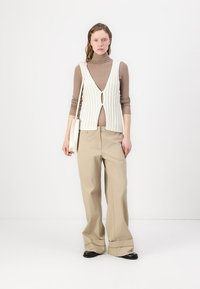 Beige weite Hosen kombiniert mit einem cremefarbenen Rippen-Top mit einem Schlüsselloch-Detail. Das Modell trägt eine kleine weiße Tasche. Neutrale Farbtöne.