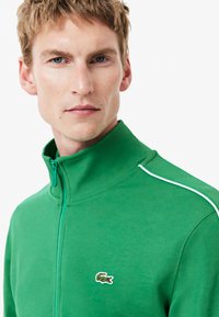 Grøn zip-up jakke lavet af blødt stof, med høj krave, hvid kontrastsyning, og et lille broderet logo på venstre side.