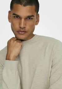 Lichtbeige crew-neck trui met ribbelmanchetten, geweven textuur en een relaxed fit. Model met hand op de kin, neutrale uitdrukking.