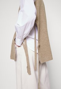 Personne portant une chemise blanche à manches longues avec un gilet en maille beige posé sur les épaules et un pantalon large de couleur claire, debout de profil.