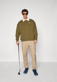Olivengrønn hettegenser med elfenbensfarget krage og broderte detaljer, parret med beige bukser og blå sko. Står med en golfkølle.