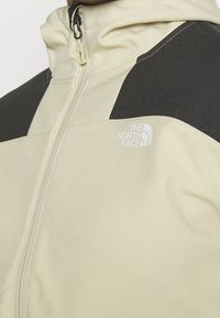 Detailní záběr béžové a černé bundy na zip s logem The North Face na prsou.