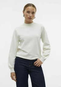 Haut à manches longues blanc avec un col montant, coupe ample, tissu texturé, poignets froncés, associé à un jean en denim sombre.