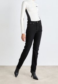 Camiseta blanca de manga larga con paneles laterales negros y media cremallera, combinada con unos jeans negros ajustados y tacones altos negros puntiagudos.